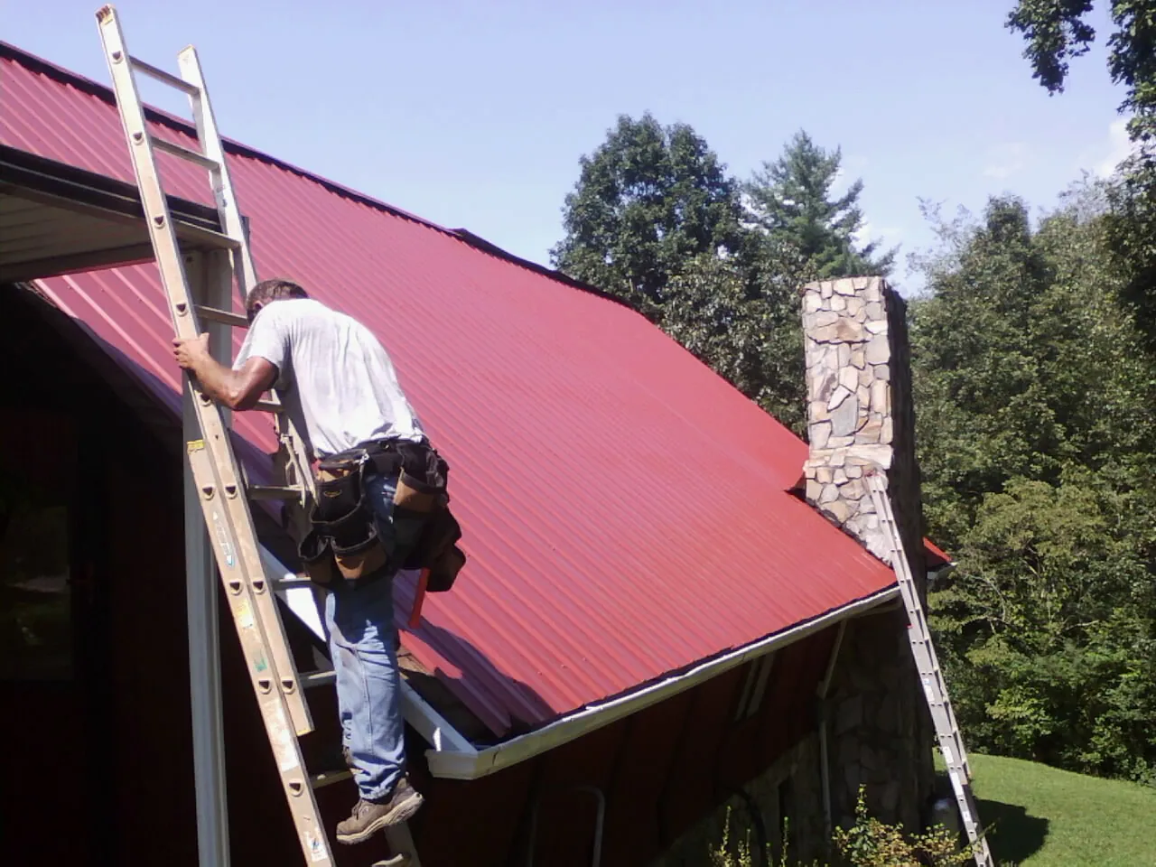 Roofer on a ladder beside a red metal roof and stone chimney — Soffit & Fascia Repair by Roofing Brothers in Suncook