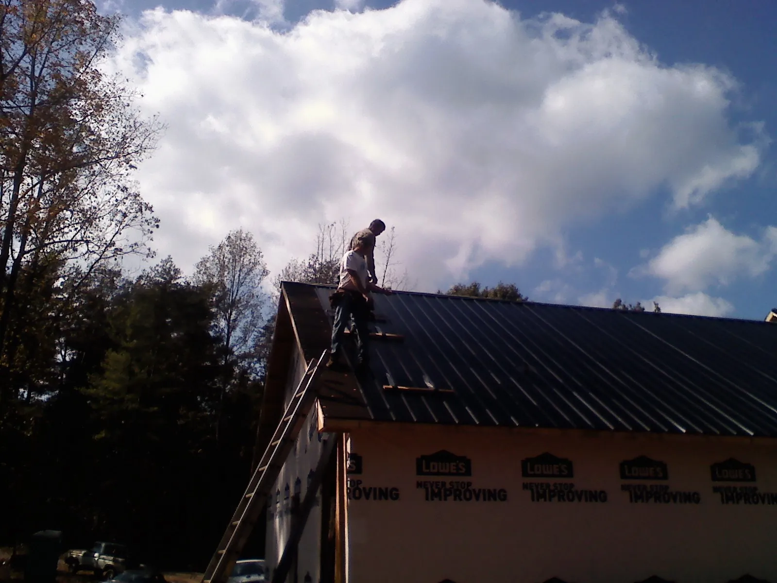 Roofer fastening dark metal panels high against a cloudy sky, from a Roof Inspection job in Suncook